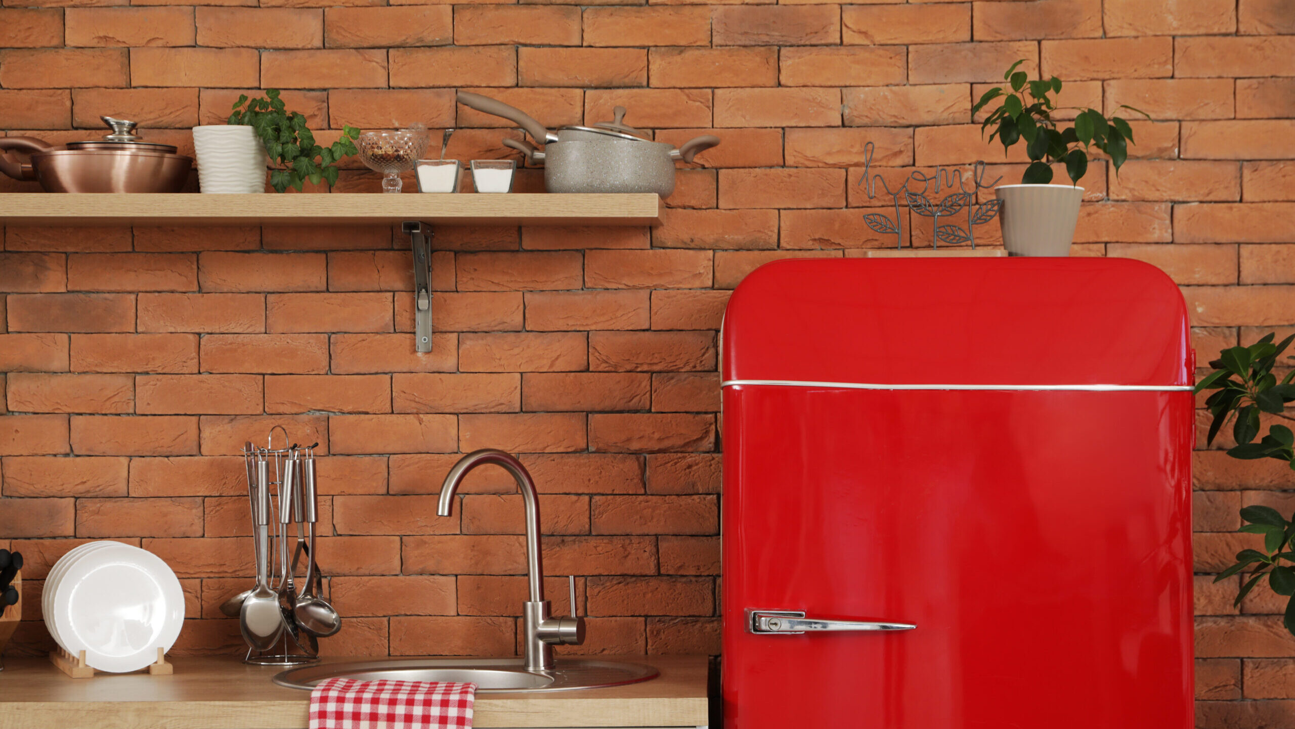 A bright red refrigerator next to a kitchen sink, with a checkered red and white towel on the counter, a wooden shelf above holding small plants and pots, and a brick wall as the backdrop, creating a cozy, modern kitchen atmosphere