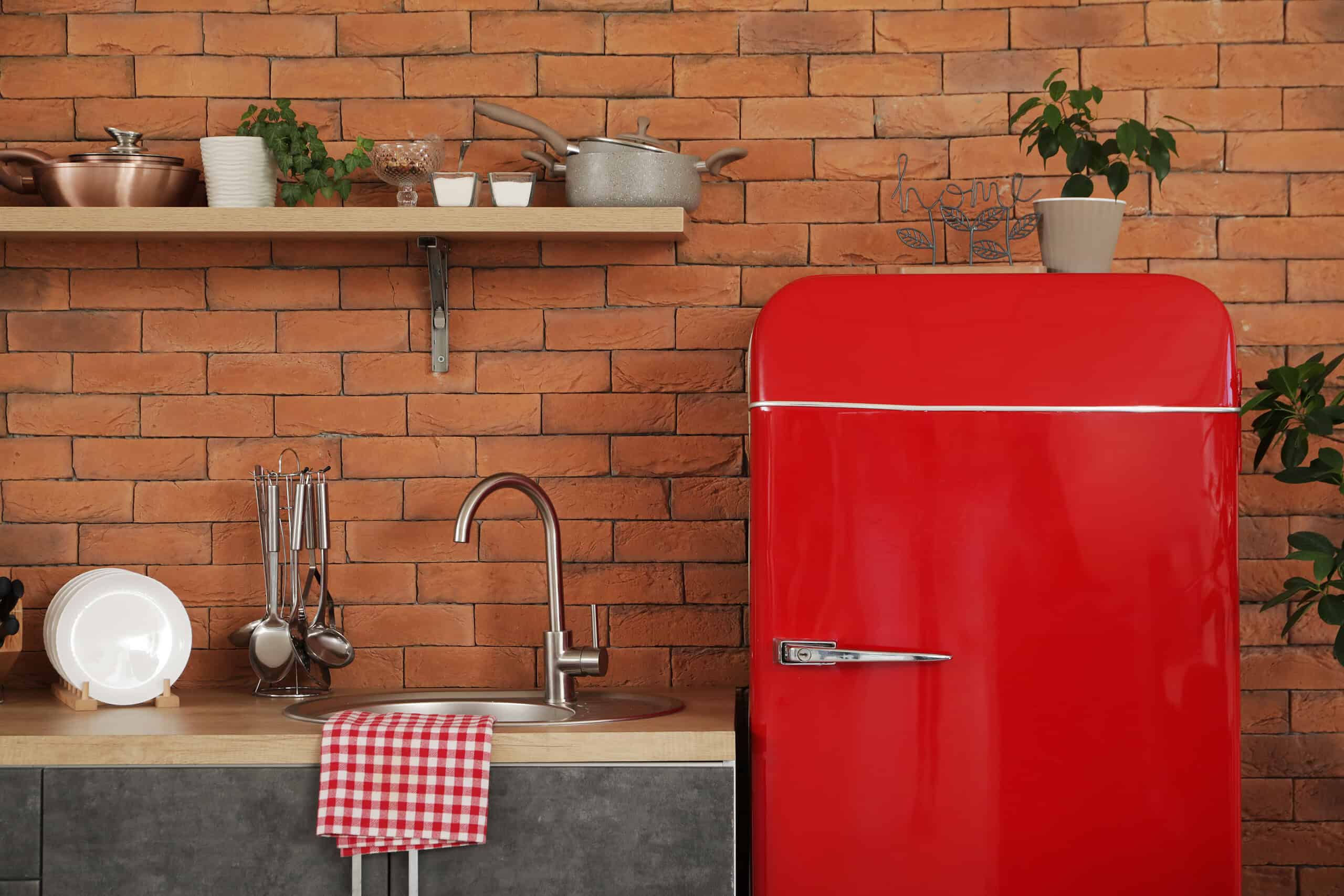 A bright red refrigerator next to a kitchen sink, with a checkered red and white towel on the counter, a wooden shelf above holding small plants and pots, and a brick wall as the backdrop, creating a cozy, modern kitchen atmosphere