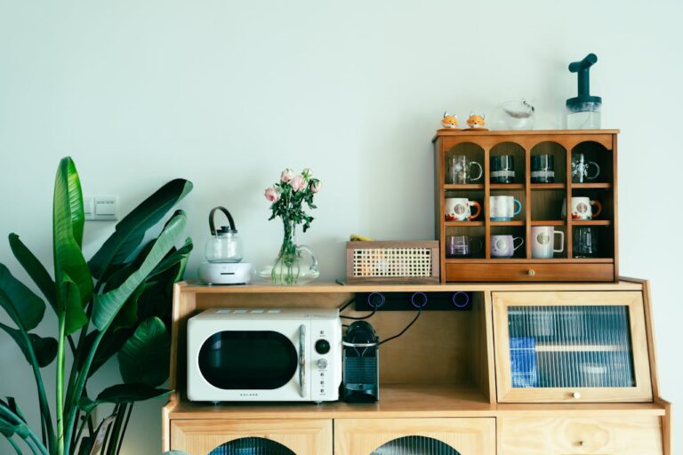 A wooden cabinet with a microwave, a coffee machine, and a shelf holding mugs, a small decorative plant on the left, a glass kettle and a vase with pink roses on the top, a beige drawer at the bottom, and a light green wall, creating a tidy and cozy kitchen corner