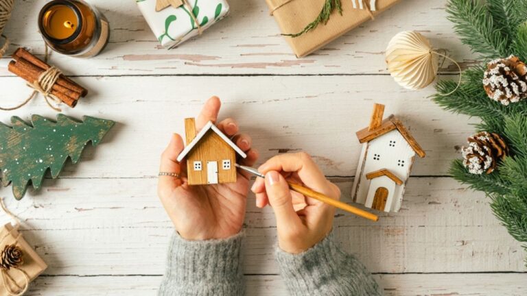 Christmas Village. Flat lay of woman hands holding brush, painting homemade wooden house on white background with Christmas natural eco decoration. Top view gifts, candle, fir tree, toy, cinnamon, pine cone, craft.