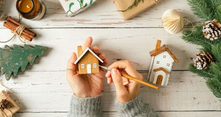Christmas Village. Flat lay of woman hands holding brush, painting homemade wooden house on white background with Christmas natural eco decoration. Top view gifts, candle, fir tree, toy, cinnamon, pine cone, craft.