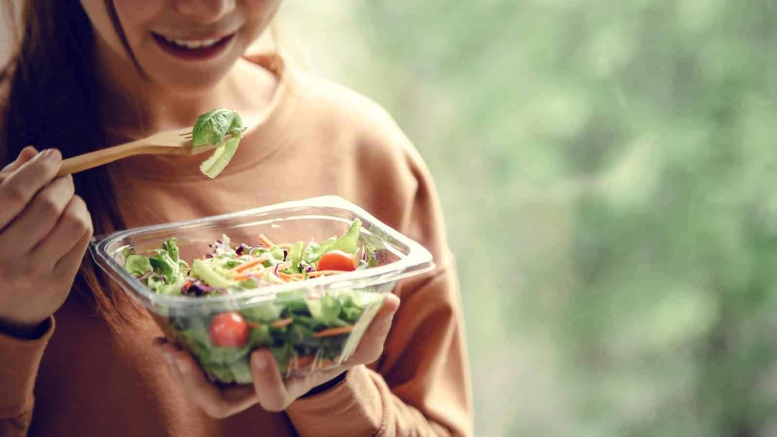 A person holding a plastic container of salad, tossing greens with one hand, vibrant fresh vegetables, casual outfit, soft background, healthy eating