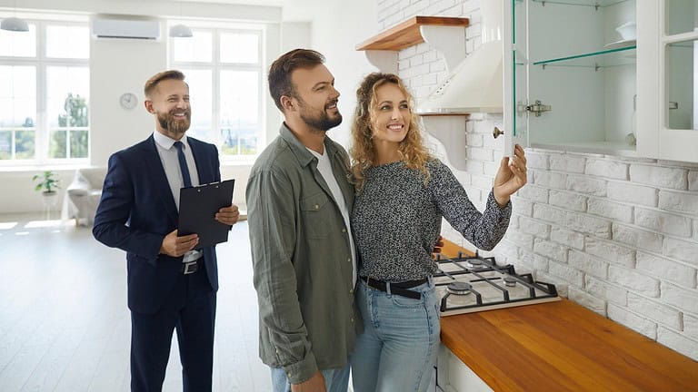 A couple exploring a kitchen during a home showing, a real estate agent in the background with a clipboard