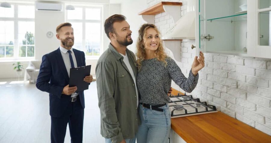 A couple exploring a kitchen during a home showing, a real estate agent in the background with a clipboard