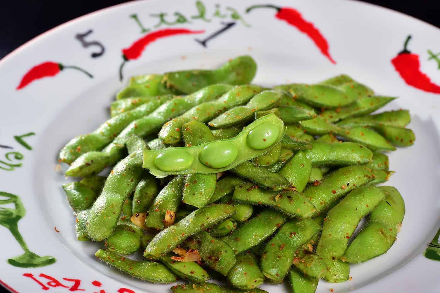 Plate of bright green edamame beans, salted and seasoned, served in pods on decorative dish with red trim