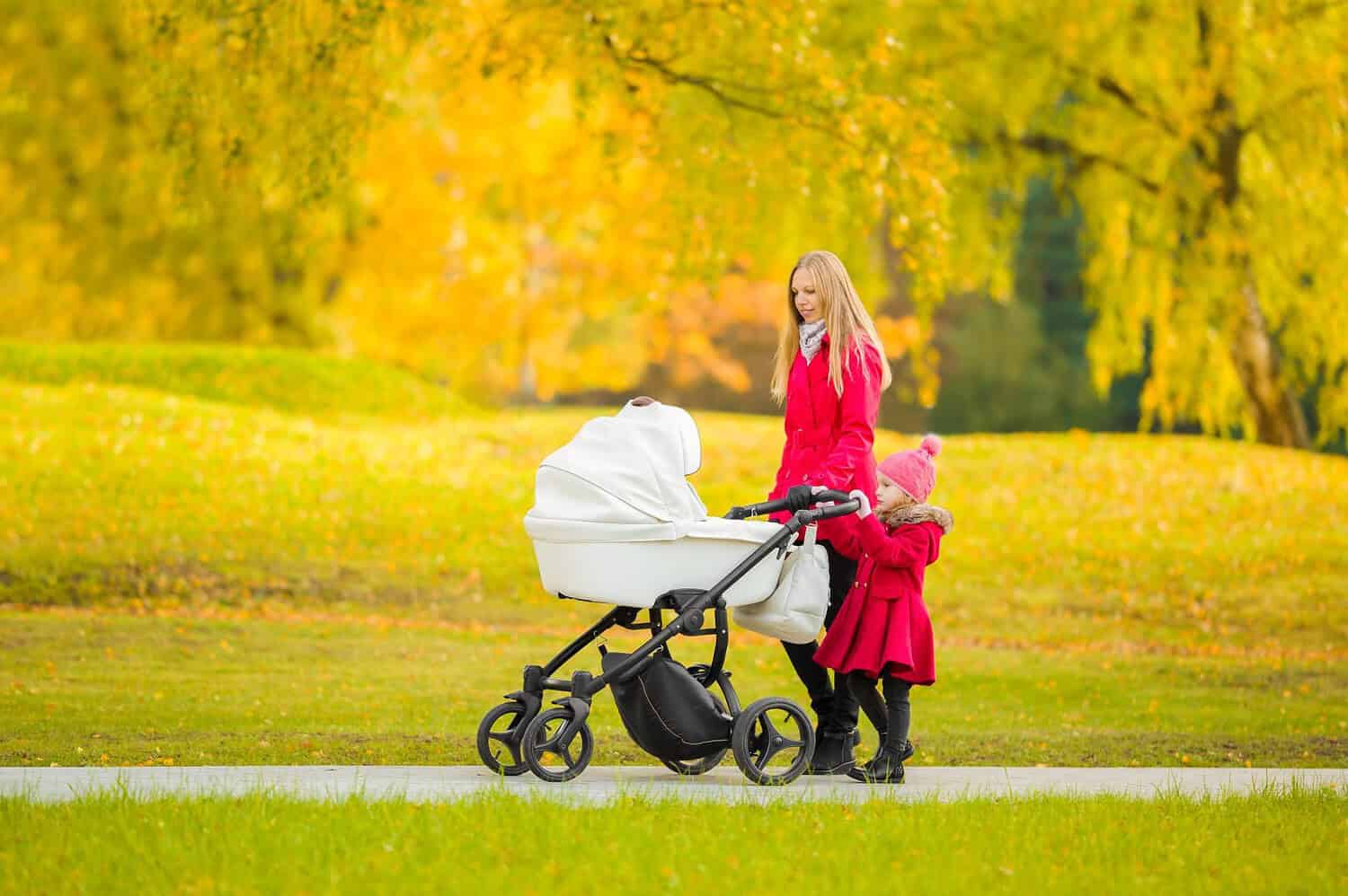 A woman pushing a stroller with a baby, accompanied by a young girl walking beside her, both wearing red coats, walking in a park with vibrant autumn foliage, sunny weather, leisurely stroll