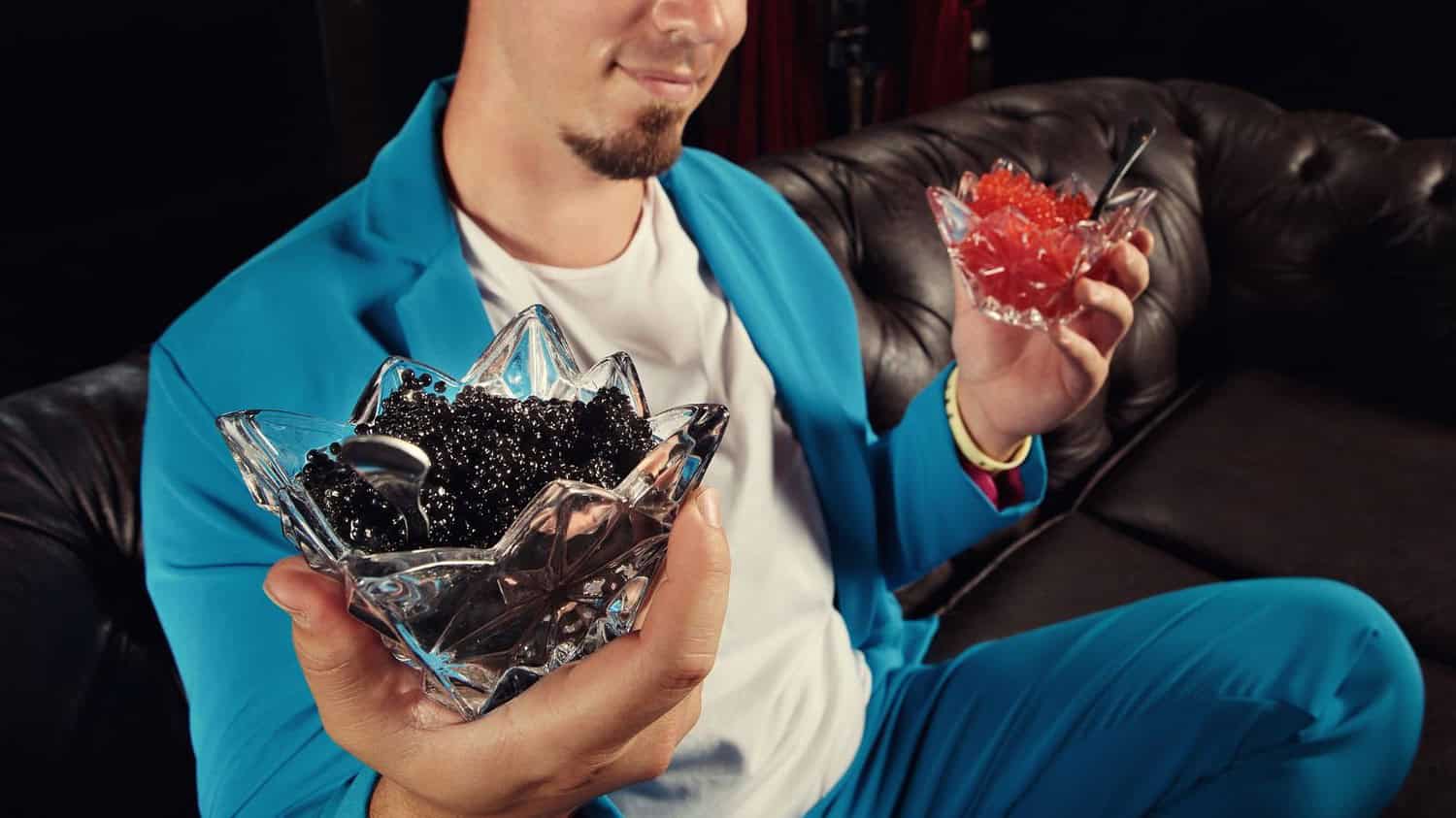 Man holding two crystal bowls filled with red and black caviar, dressed in a blue suit, elegant presentation of luxury food, person displaying gourmet delicacies