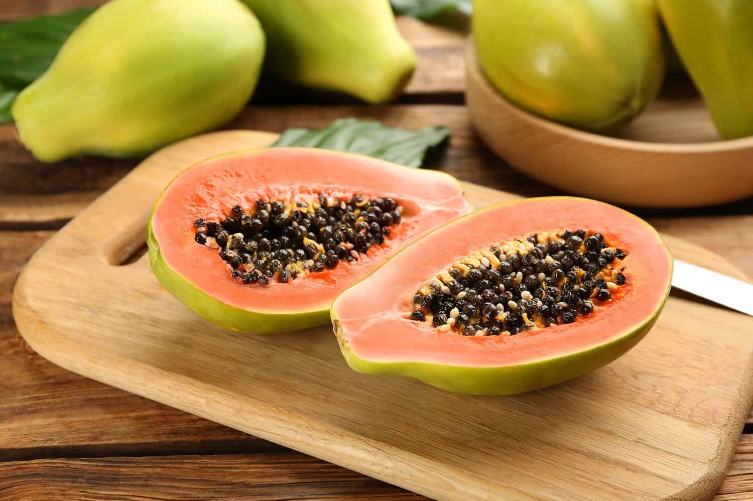 Papaya cut in half, showing orange flesh, black seeds in the center, placed on a wooden cutting board, fresh and ripe fruit, green papaya in the background