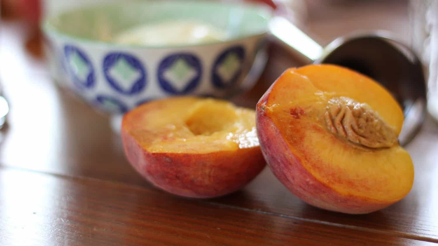 Fresh peach cut in half showing orange flesh and brown pit, placed on wooden surface near patterned cup