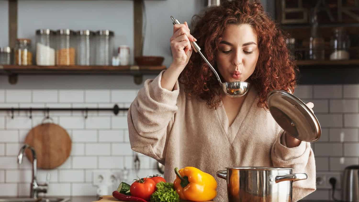 A woman in a kitchen, holding a ladle, tasting a soup from a pot, fresh vegetables like tomatoes and bell peppers are visible on the counter, casual cooking at home, preparing a meal