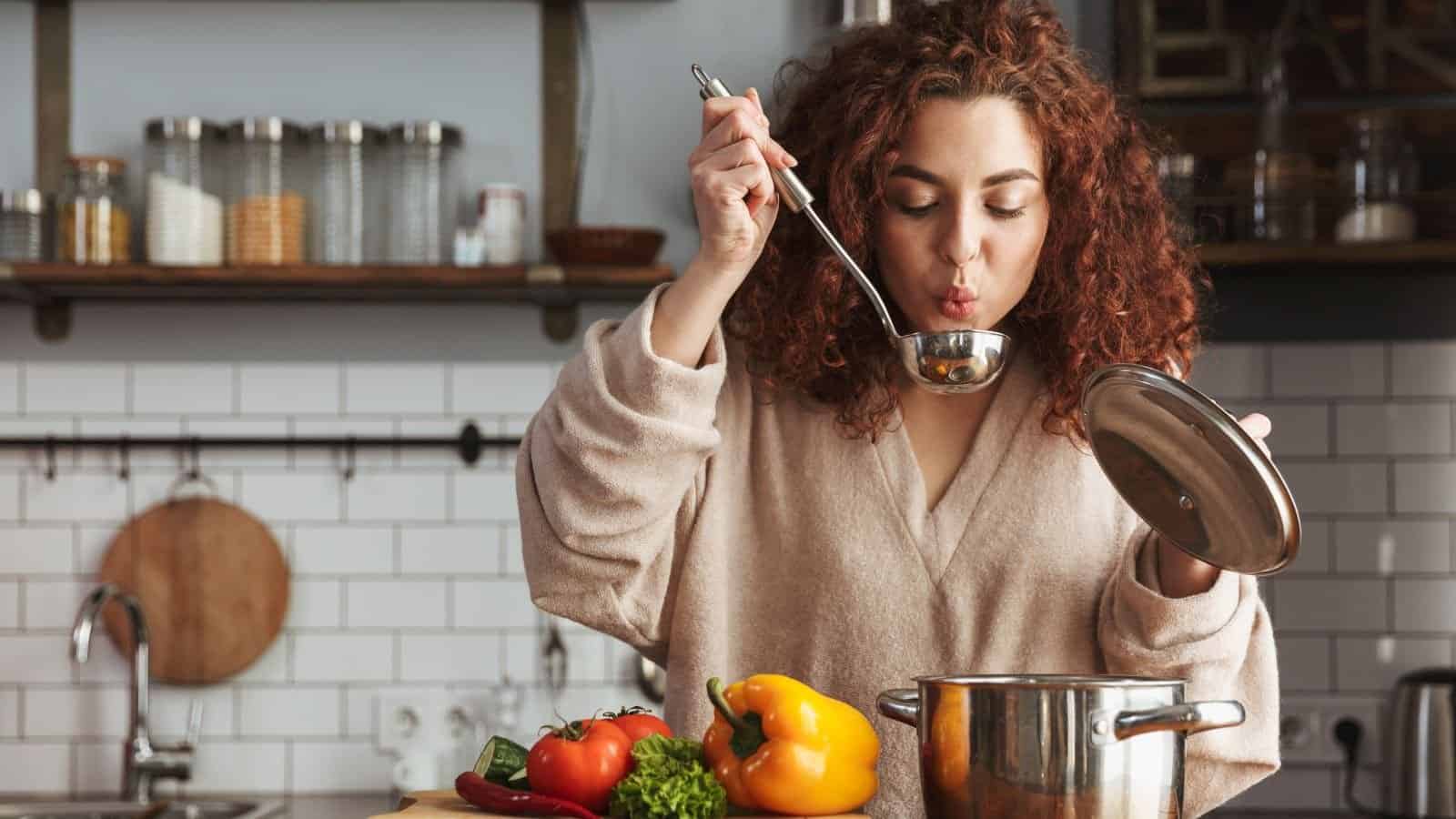 A woman in a kitchen, holding a ladle, tasting a soup from a pot, fresh vegetables like tomatoes and bell peppers are visible on the counter, casual cooking at home, preparing a meal