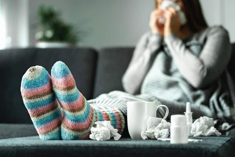 Person with cold or flu wearing striped socks, sitting on couch with tissues and hot drink nearby