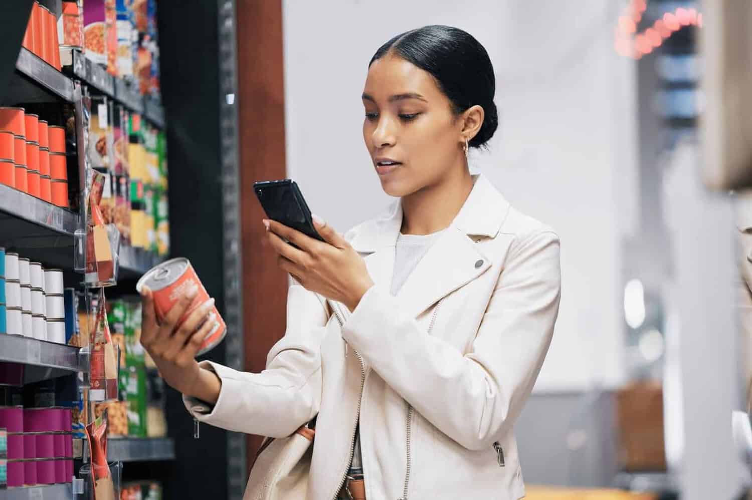 Woman in a grocery store, holding a can in one hand, using a smartphone with the other, checking product information or prices, looking at the product label