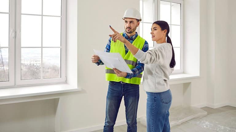 Meeting female homeowner and builder in living room for further renovation work.Attentive foreman in helmet and overalls with document or technical plan in his hands communicates with hostess of house