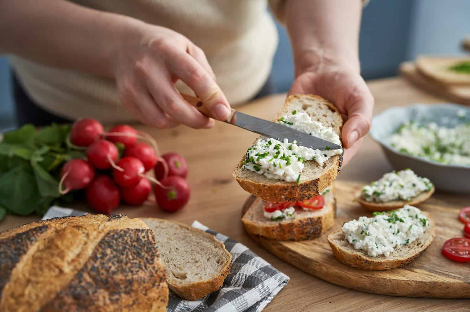 Person spreading herb-infused cream cheese on rustic bread, fresh radishes on the table, homemade sandwich preparation, wooden cutting board, cozy kitchen setting, healthy and fresh ingredients