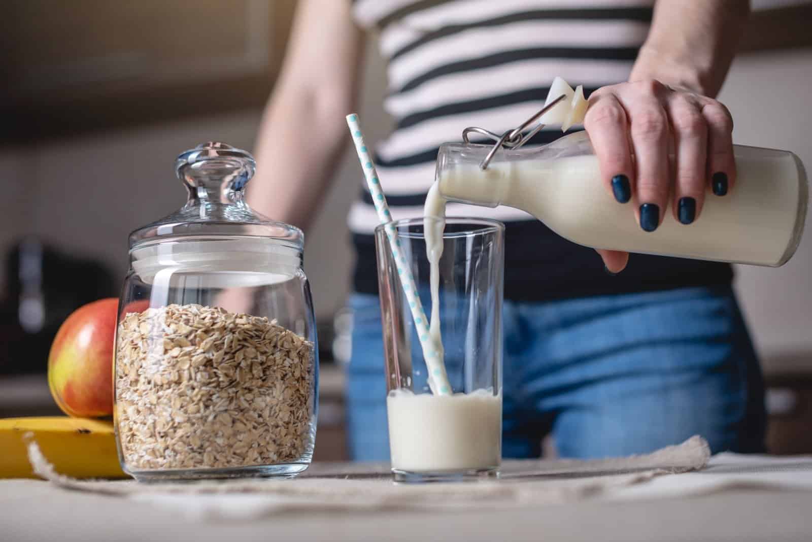 Person in striped shirt pouring milk into glass, cereal jar and bowl on counter, kitchen breakfast preparation scene