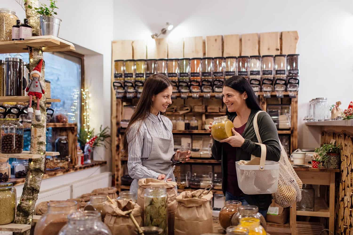Two women shopping at a bulk store, one holding a jar of liquid, the other holding a glass, wooden shelving filled with jars of grains and spices in the background