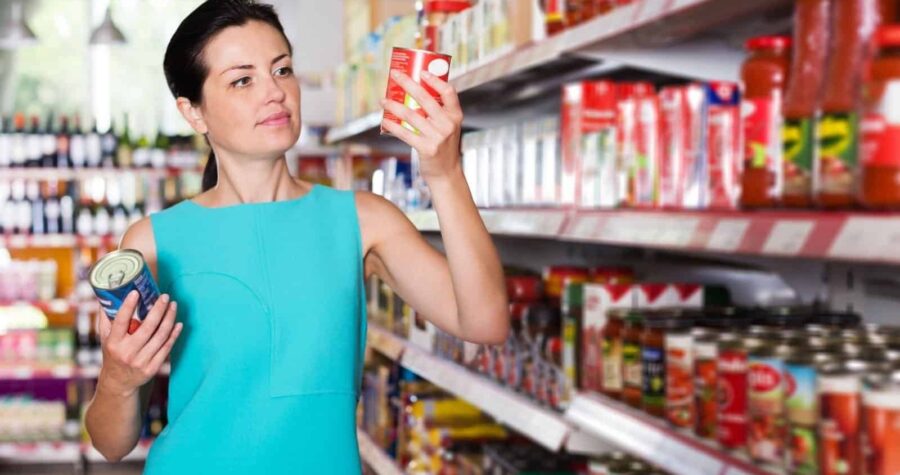 A woman in a teal dress examines a can, grocery store aisle, shelves filled with various canned goods, focused on reading product label, making a purchase decision, shopping for groceries, in a well-lit supermarket