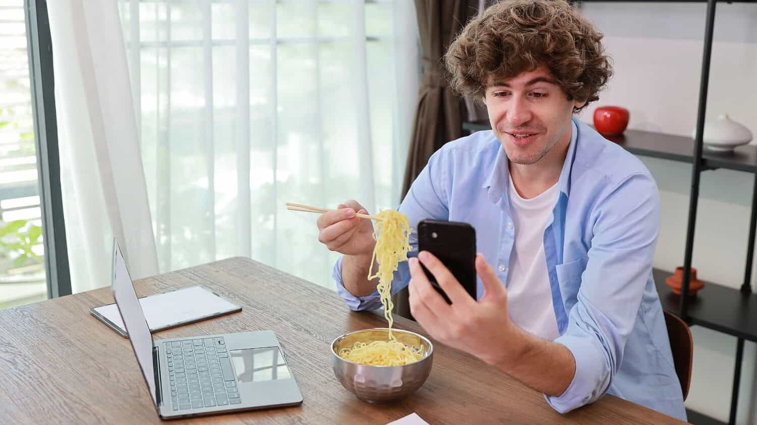 Man eating noodles, holding chopsticks, looking at phone, smiling, sitting at a table, working or relaxing, laptop and bowl of noodles in front