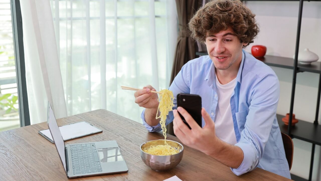 Man eating noodles, holding chopsticks, looking at phone, smiling, sitting at a table, working or relaxing, laptop and bowl of noodles in front