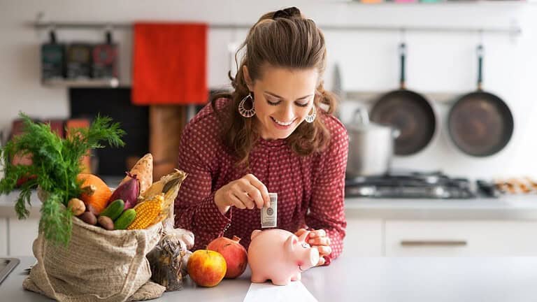 Smiling woman in a kitchen placing money into a pink piggy bank beside a bag of fresh vegetables and fruits