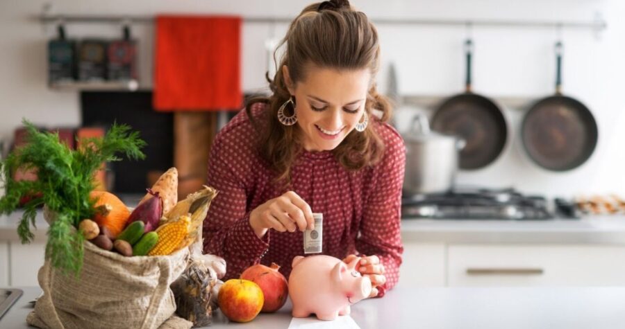 Smiling woman in a kitchen placing money into a pink piggy bank beside a bag of fresh vegetables and fruits