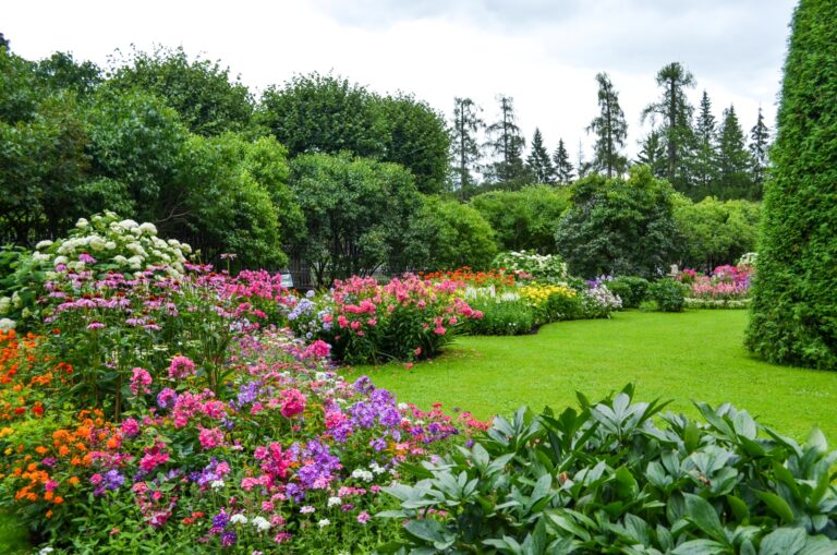 Manicured garden with vibrant flower beds, green lawn, mature trees, and colorful blooms in landscaped park setting
