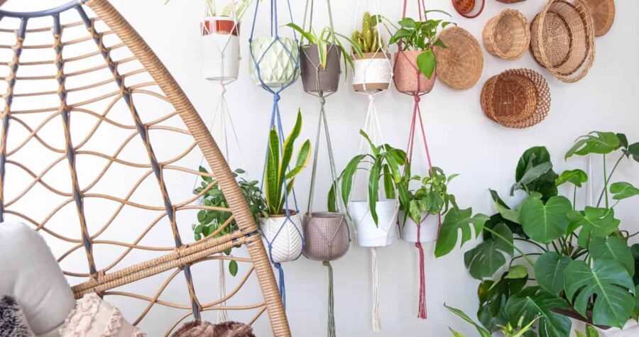 Multiple macrame plant hangers with indoor houseplants and pot planters are hanging from a metal pole. Boho basket wall decor and wicker egg chair are use to add character to the cozy bohemian room.