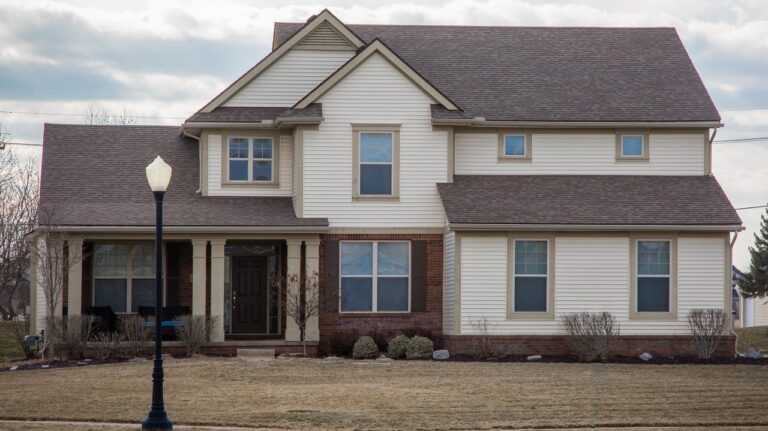 A traditional two-story family home in a middle class neighborhood looks outdated with off-white vinyl siding, brick, simple beige window frames with no shutters and a brown shingles roof.