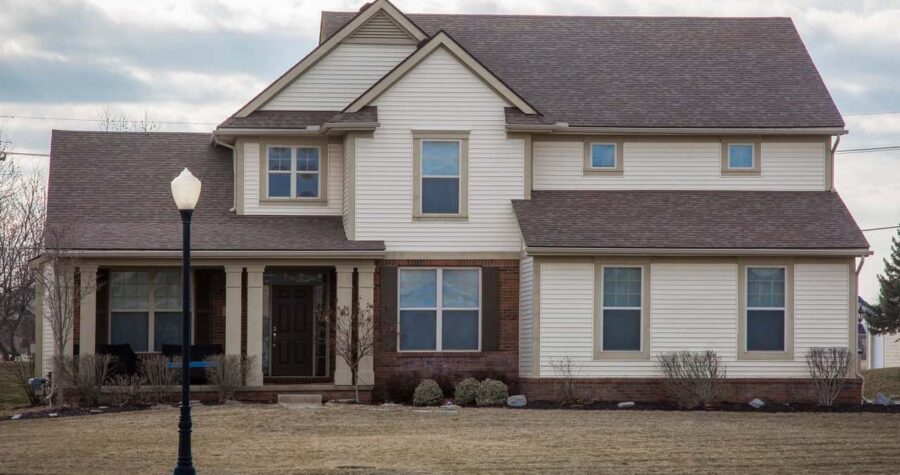 A traditional two-story family home in a middle class neighborhood looks outdated with off-white vinyl siding, brick, simple beige window frames with no shutters and a brown shingles roof.
