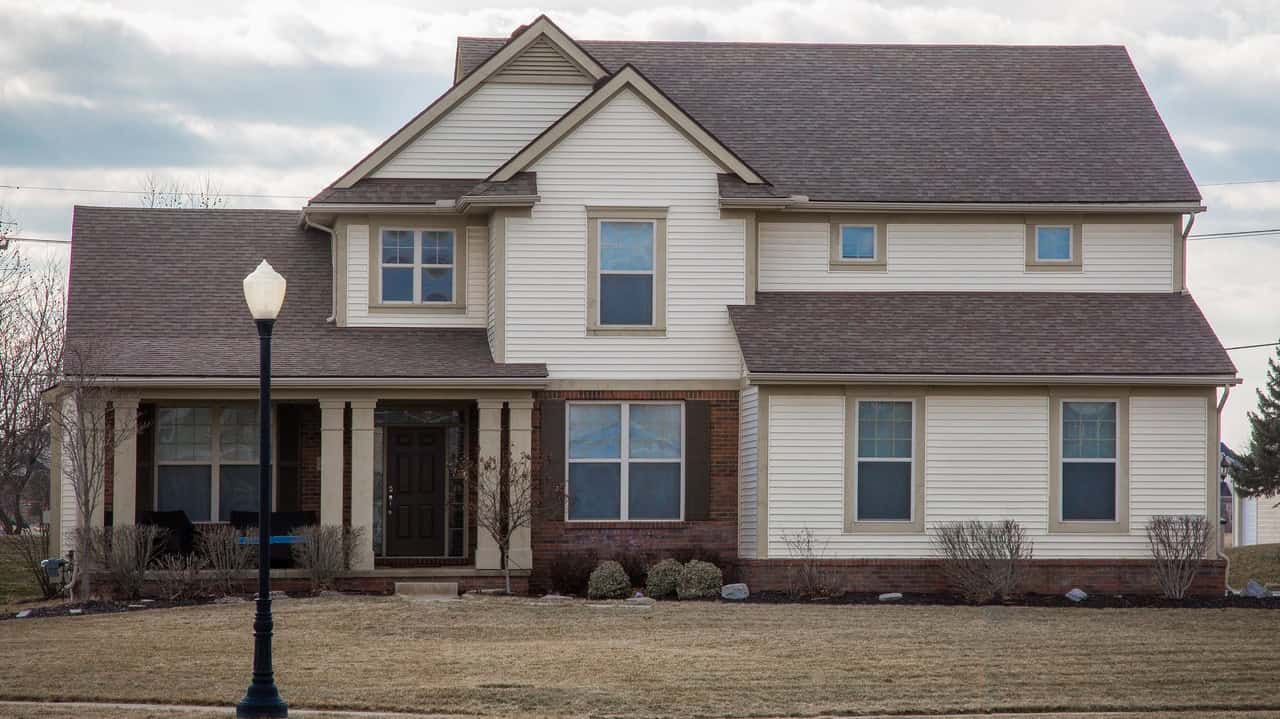 A traditional two-story family home in a middle class neighborhood looks outdated with off-white vinyl siding, brick, simple beige window frames with no shutters and a brown shingles roof.