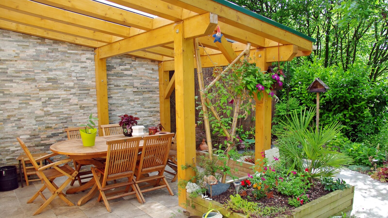Outdoor patio with a wooden pergola, dining table with chairs, potted plants on the floor, colorful hanging plants, garden beds with flowers, stone flooring, cozy and inviting setting