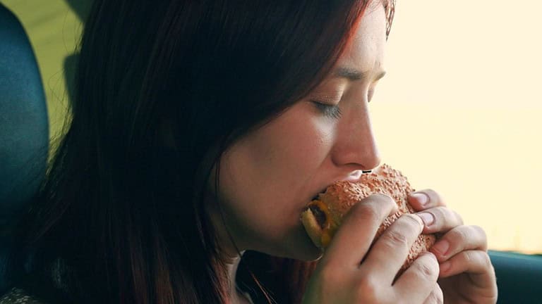 Girl passenger eats burger in car. Delicious food bun with cutlet. Young hungry woman eats roll with meatball while sitting in car. Teenager travels, having snack on road without leaving car. Tourist