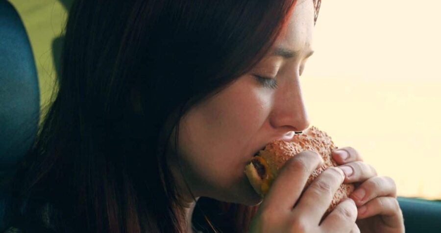 Girl passenger eats burger in car. Delicious food bun with cutlet. Young hungry woman eats roll with meatball while sitting in car. Teenager travels, having snack on road without leaving car. Tourist
