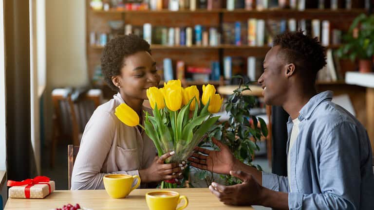 Handsome black guy giving his girlfriend bouquet of tulips and gift for Valentine's Day at cafe. Affectionate African American couple celebrating holiday, having romantic moments at coffee shop