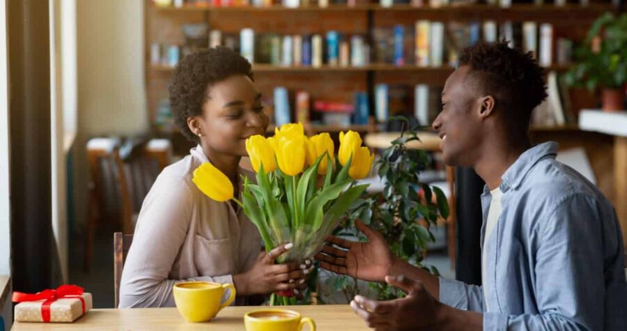 Handsome black guy giving his girlfriend bouquet of tulips and gift for Valentine's Day at cafe. Affectionate African American couple celebrating holiday, having romantic moments at coffee shop