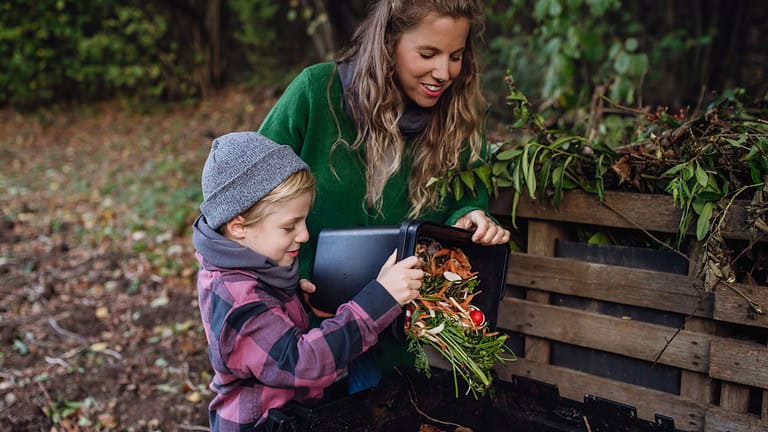 Boy helping mother to put kitchen waste, peel and leftover vegetables scraps into composter in the garden