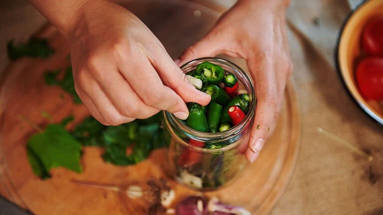 Hands placing chili peppers into a jar, close-up of jarred vegetables, adding peppers to a glass jar for storage