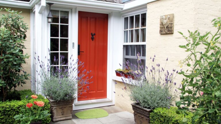 Red wooden front door, white-framed windows, potted lavender plants, green doormat, beige brick wall, cozy home entrance