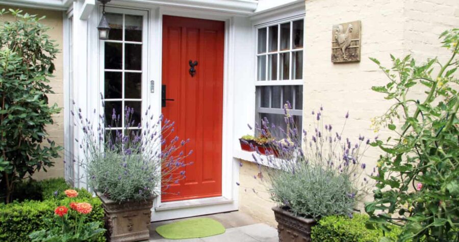 Red wooden front door, white-framed windows, potted lavender plants, green doormat, beige brick wall, cozy home entrance