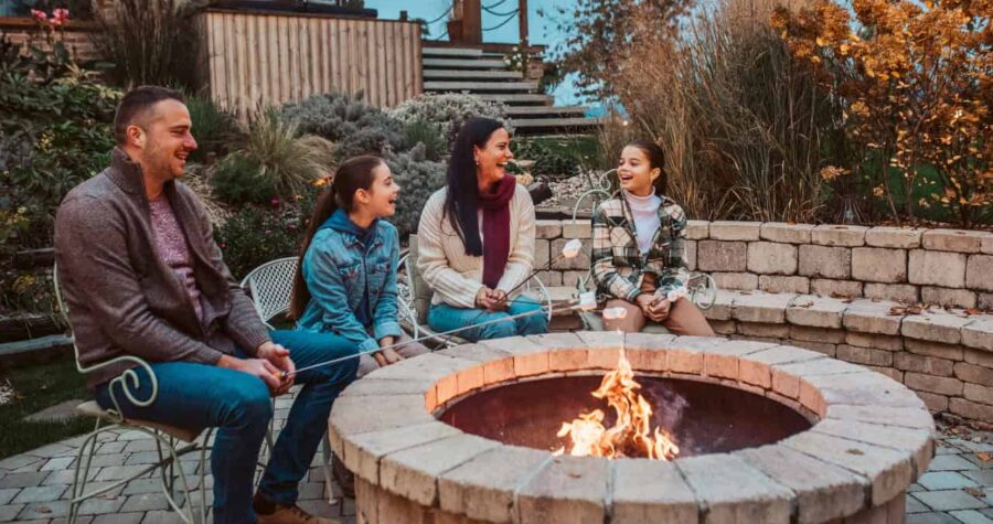 Family of four sitting around outdoor stone fire pit in backyard during evening, with flames burning in center