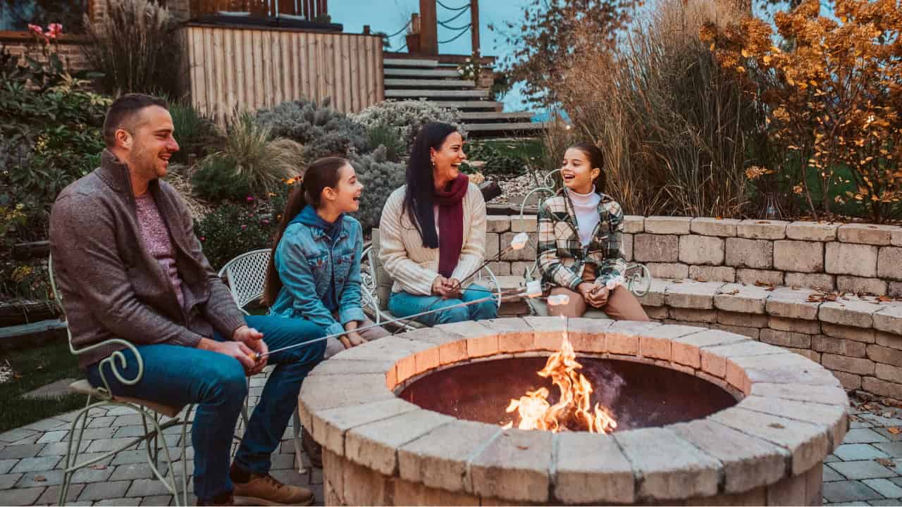 Family of four sitting around outdoor stone fire pit in backyard during evening, with flames burning in center