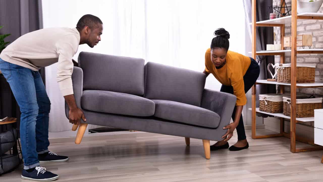 Side View Of A Smiling Young African Couple Lifting Sofa In Living Room