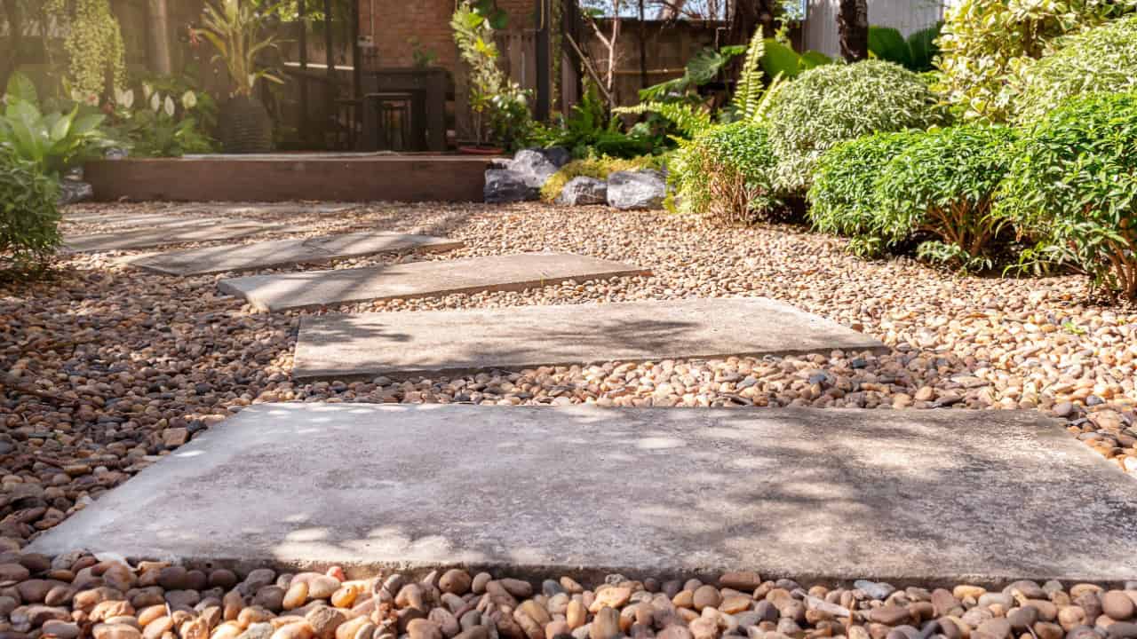 A garden pathway covered in small gravel, featuring large concrete slabs, surrounded by greenery