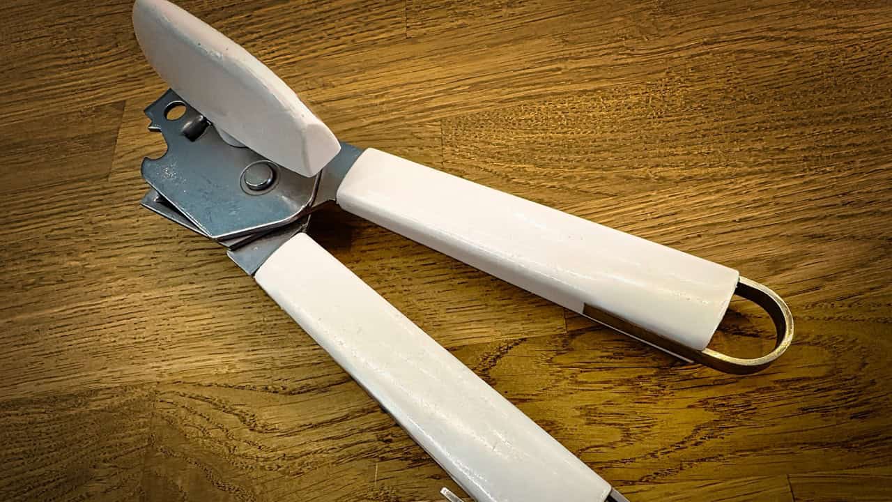 The image shows a manual can opener, with white handles and a metal blade for opening cans, placed on a wooden surface