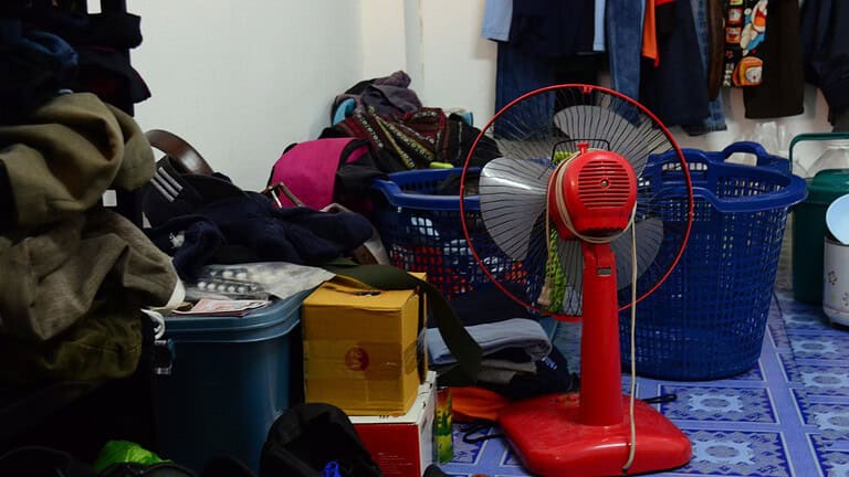 Cluttered room, red standing fan, laundry baskets with clothes, plastic storage bins, scattered bags, stacked boxes