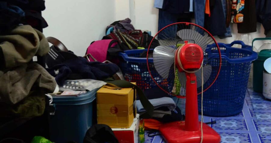 Cluttered room, red standing fan, laundry baskets with clothes, plastic storage bins, scattered bags, stacked boxes