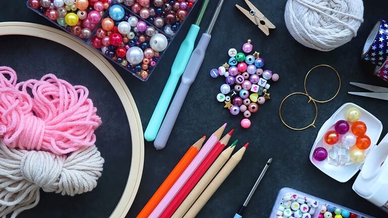 Assorted crafting supplies including colorful beads, crochet hooks, colored pencils, embroidery hoop, and sewing tools, arranged on a dark surface, ideal for DIY projects and creative hobbies