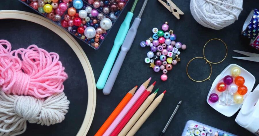 Assorted crafting supplies including colorful beads, crochet hooks, colored pencils, embroidery hoop, and sewing tools, arranged on a dark surface, ideal for DIY projects and creative hobbies