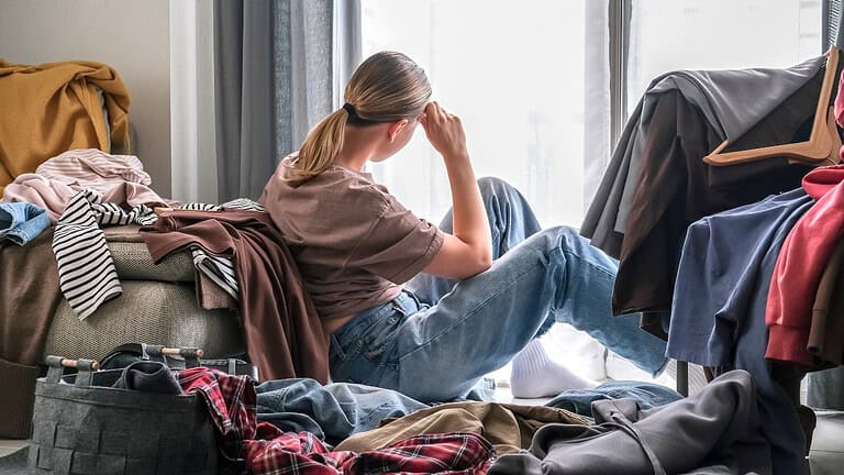 A person sitting among a pile of clothes, feeling overwhelmed, looking out the window, disorganized room with clothes scattered on the floor and hanging